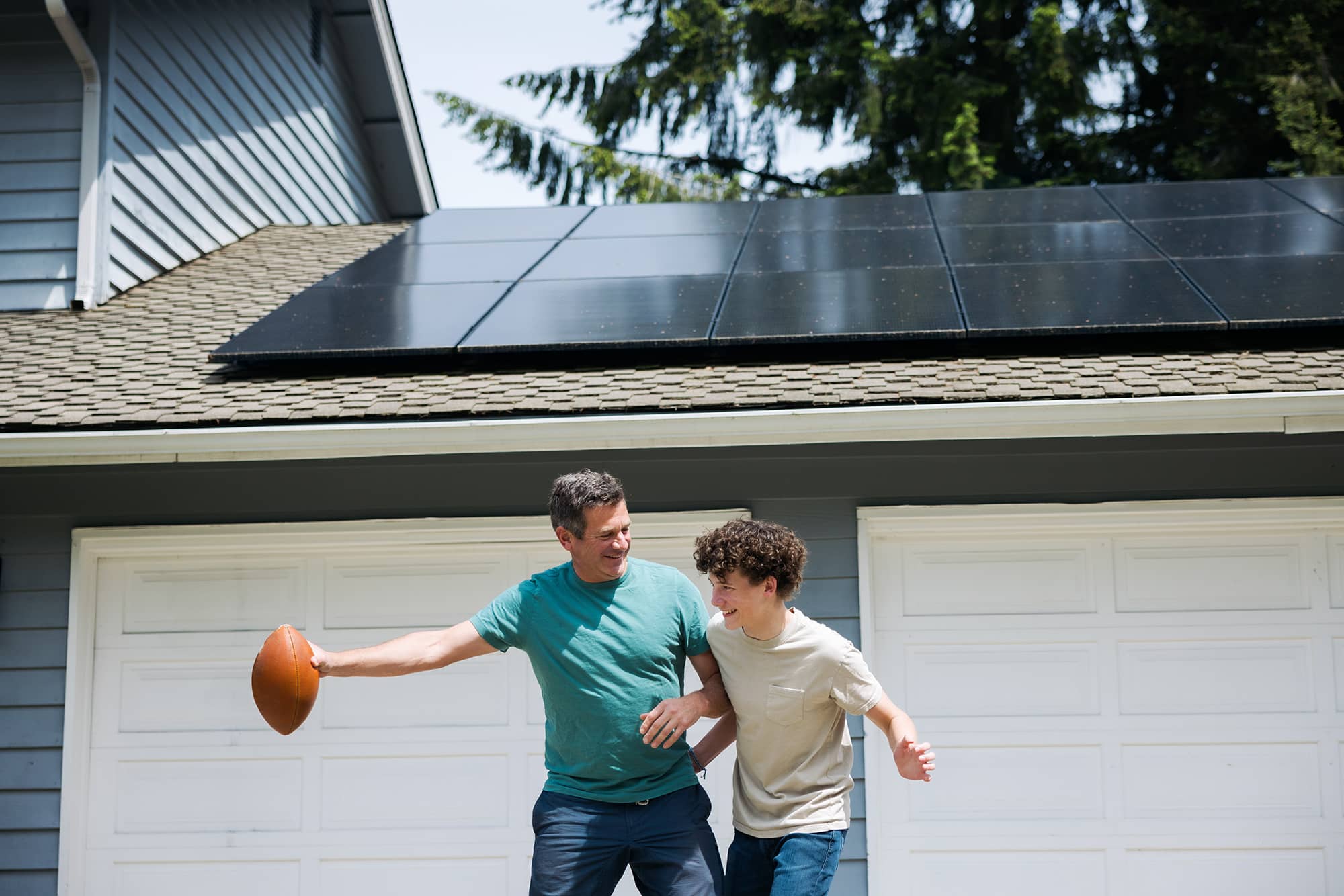 A man and a teenage boy play with an American football in front of a house with solar panels on the roof and white garage doors. The man is smiling as he holds the football, while the boy tries to block him.